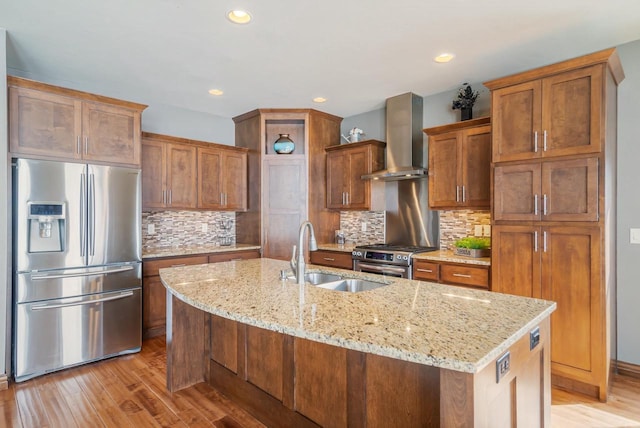 kitchen featuring wall chimney range hood, sink, a kitchen island with sink, stainless steel appliances, and light stone counters