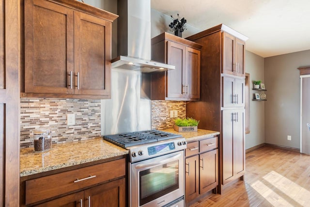 kitchen with stainless steel gas range, backsplash, light stone counters, wall chimney exhaust hood, and light wood-type flooring