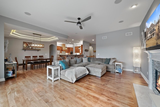 living room with ceiling fan, a tray ceiling, a stone fireplace, and light wood-type flooring