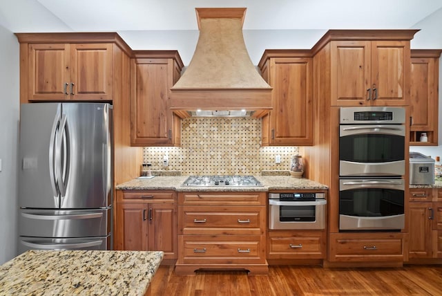 kitchen with custom range hood, stainless steel appliances, light stone countertops, hardwood / wood-style floors, and backsplash