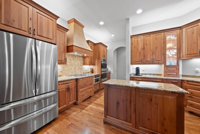 kitchen featuring stainless steel appliances, light stone counters, custom range hood, a kitchen island, and light wood-type flooring