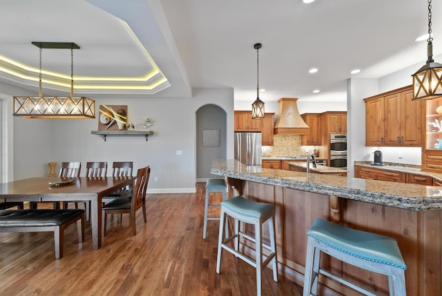 kitchen with custom exhaust hood, a breakfast bar area, hanging light fixtures, appliances with stainless steel finishes, and a raised ceiling