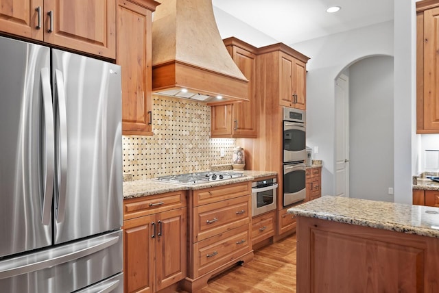 kitchen with decorative backsplash, light stone counters, stainless steel appliances, custom range hood, and light hardwood / wood-style flooring