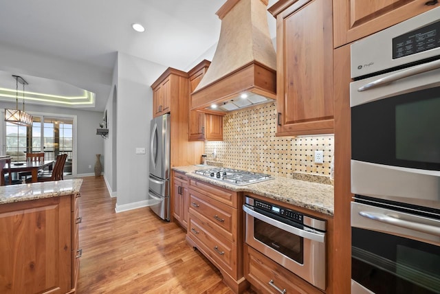 kitchen with premium range hood, appliances with stainless steel finishes, tasteful backsplash, light stone counters, and a tray ceiling