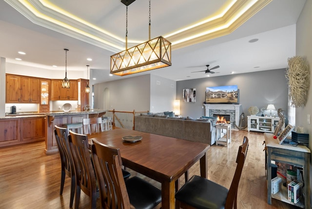 dining space with ceiling fan, a stone fireplace, a raised ceiling, and light hardwood / wood-style flooring