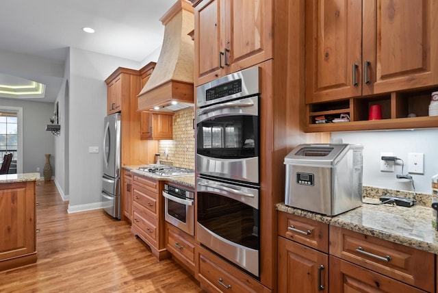 kitchen featuring premium range hood, appliances with stainless steel finishes, light stone counters, decorative backsplash, and light wood-type flooring