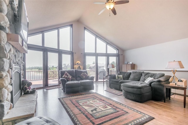 living room featuring a stone fireplace, light hardwood / wood-style flooring, high vaulted ceiling, and ceiling fan