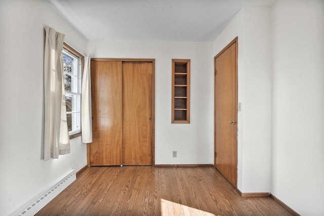 unfurnished bedroom with a baseboard heating unit, wood finished floors, baseboards, and a closet