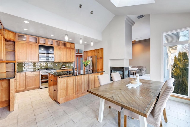 kitchen featuring dark stone counters, a multi sided fireplace, pendant lighting, stainless steel appliances, and decorative backsplash