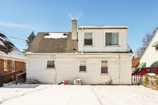 snow covered house featuring fence, a chimney, and roof with shingles