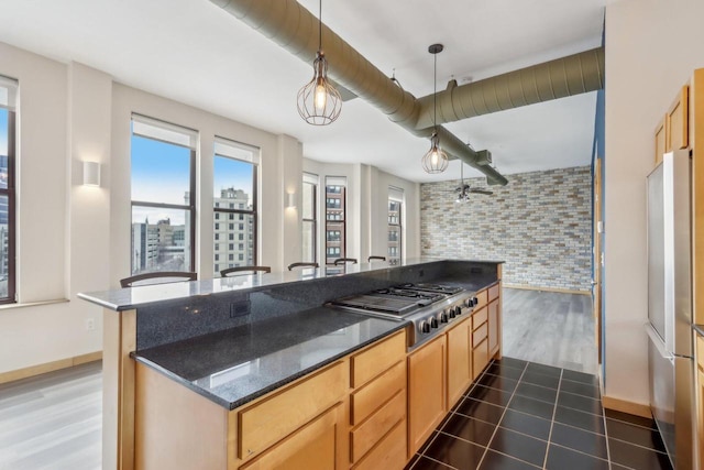 kitchen featuring stainless steel appliances, light brown cabinets, dark stone countertops, pendant lighting, and brick wall