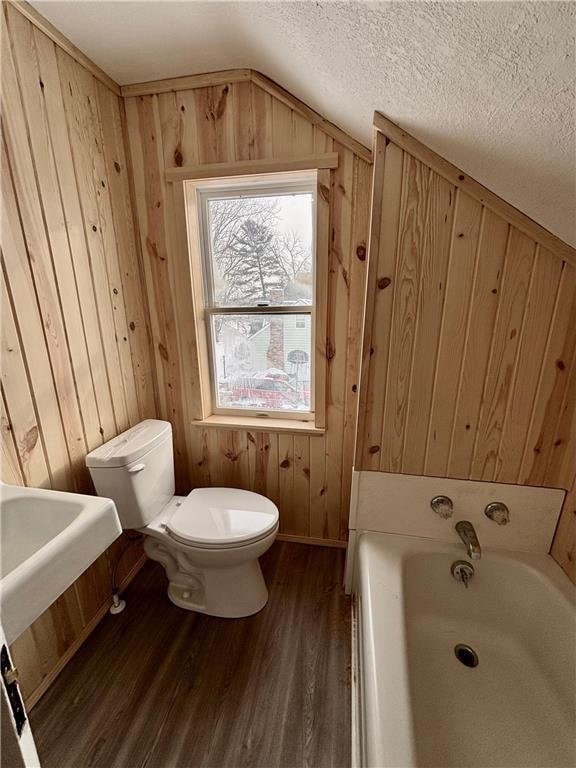 bathroom featuring hardwood / wood-style flooring, wooden walls, a bath, a textured ceiling, and toilet