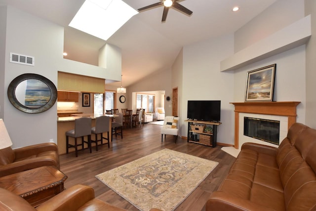 living room with ceiling fan, a tiled fireplace, a skylight, high vaulted ceiling, and dark hardwood / wood-style flooring