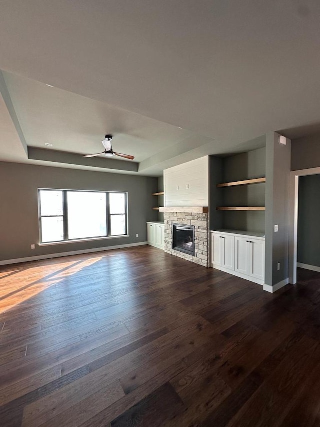 unfurnished living room featuring a stone fireplace, ceiling fan, a tray ceiling, dark wood-type flooring, and built in shelves