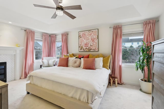 bedroom featuring light carpet, a tray ceiling, a fireplace, and ceiling fan