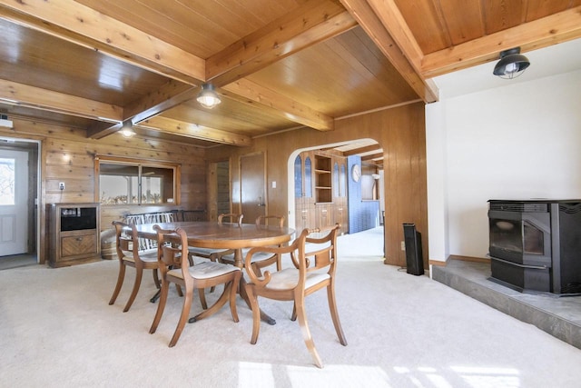 dining space featuring arched walkways, wooden walls, light carpet, beamed ceiling, and a wood stove