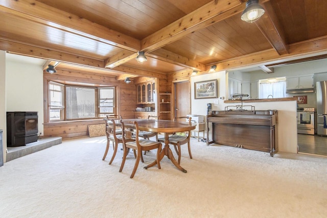 dining space featuring wooden walls, light colored carpet, wood ceiling, beamed ceiling, and a wood stove