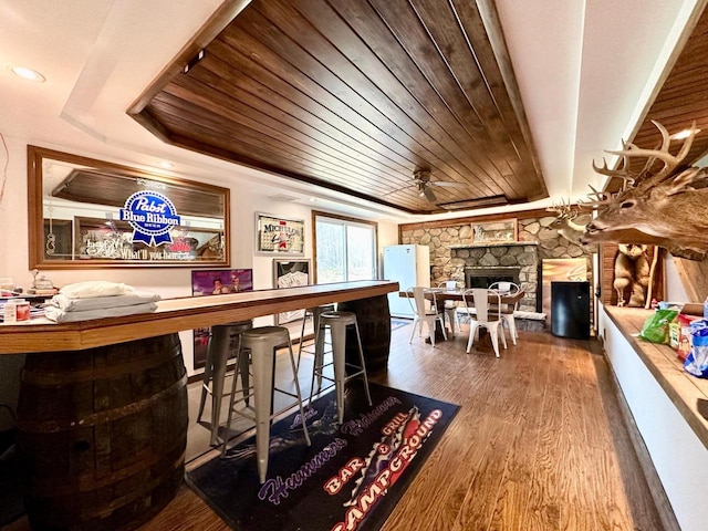 bar with wood ceiling, hardwood / wood-style flooring, ceiling fan, a stone fireplace, and a raised ceiling