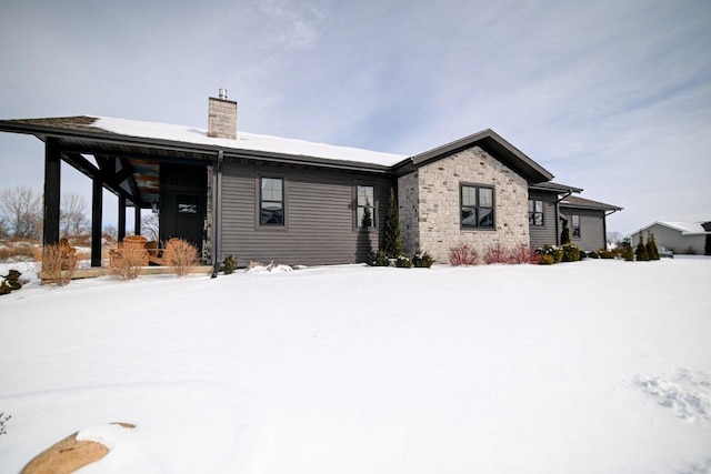 snow covered rear of property featuring stone siding and a chimney