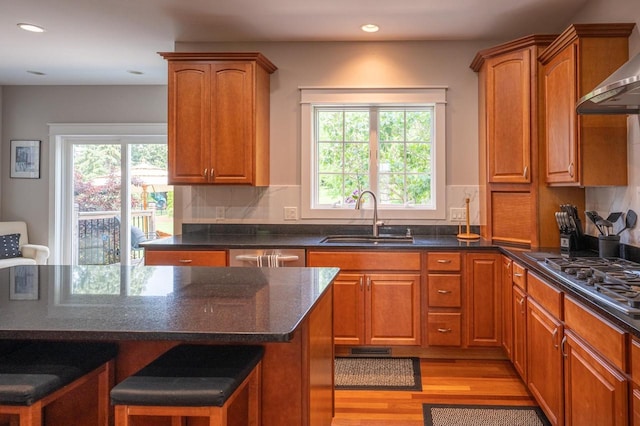 kitchen with sink, wall chimney range hood, stainless steel appliances, light hardwood / wood-style floors, and dark stone counters