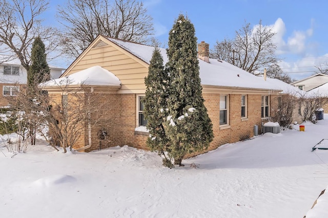 snow covered property with brick siding, a chimney, and central air condition unit