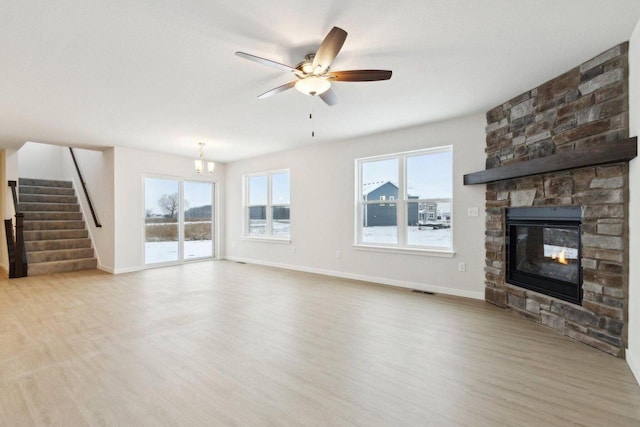 unfurnished living room with light wood-type flooring, a wealth of natural light, a stone fireplace, and ceiling fan with notable chandelier