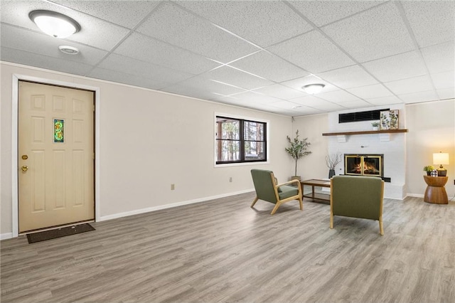 interior space with a brick fireplace, a drop ceiling, baseboards, and wood finished floors