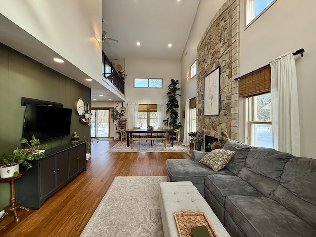 living room with recessed lighting, a towering ceiling, a ceiling fan, a stone fireplace, and wood finished floors