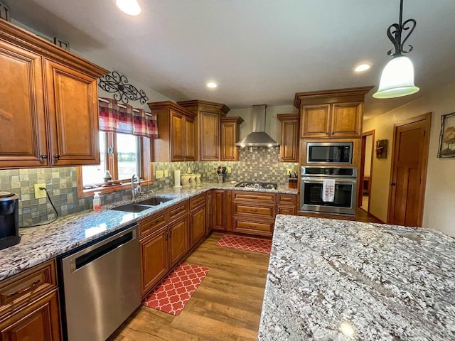 kitchen featuring a sink, appliances with stainless steel finishes, wall chimney range hood, brown cabinetry, and decorative light fixtures