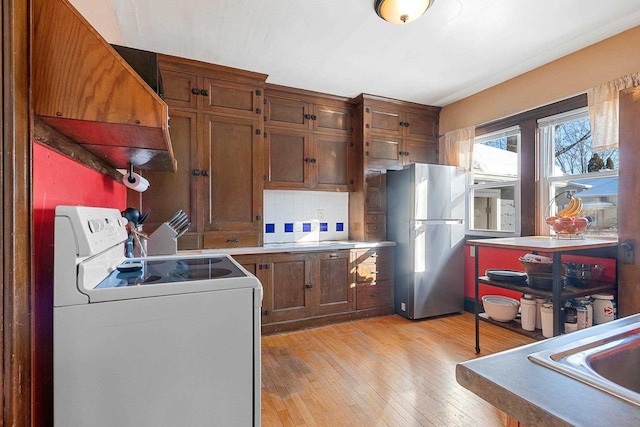 kitchen with light wood-style flooring, backsplash, white range with electric stovetop, freestanding refrigerator, and brown cabinets