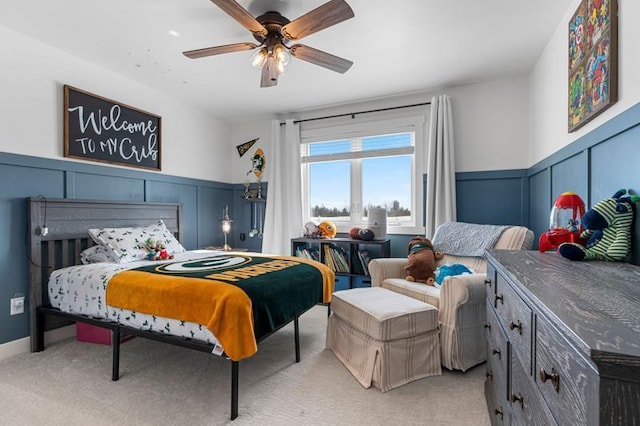 bedroom featuring a ceiling fan, light colored carpet, a wainscoted wall, and a decorative wall