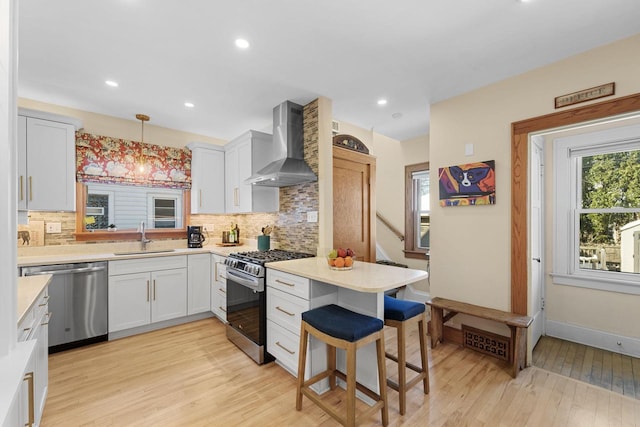 kitchen with pendant lighting, light countertops, appliances with stainless steel finishes, a sink, and wall chimney exhaust hood