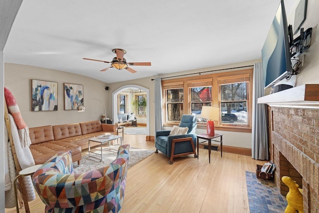 living room featuring arched walkways, a fireplace, a ceiling fan, baseboards, and light wood finished floors