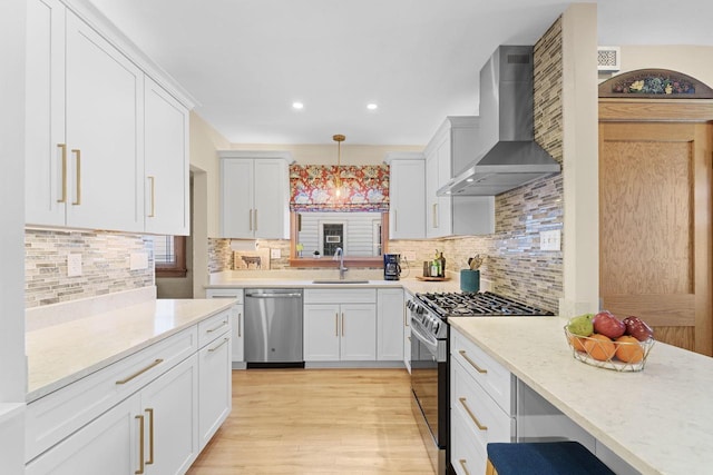 kitchen featuring pendant lighting, appliances with stainless steel finishes, white cabinets, a sink, and wall chimney exhaust hood