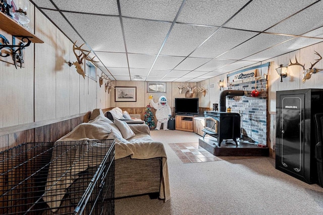 carpeted living area featuring a wood stove, wood walls, and a drop ceiling