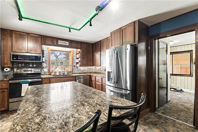 kitchen with stainless steel appliances, decorative backsplash, stone finish floor, a sink, and dark stone countertops