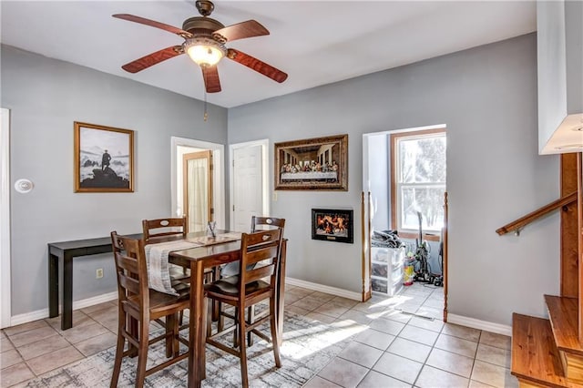 dining space with ceiling fan, a lit fireplace, baseboards, and light tile patterned floors