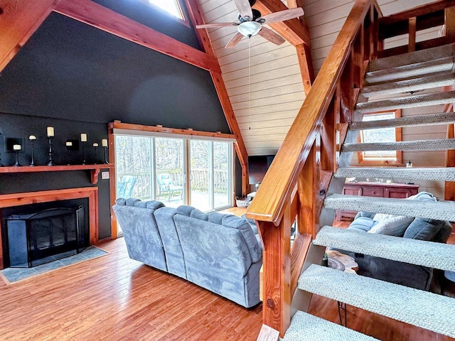 living area with stairway, lofted ceiling with beams, a fireplace with flush hearth, ceiling fan, and wood finished floors