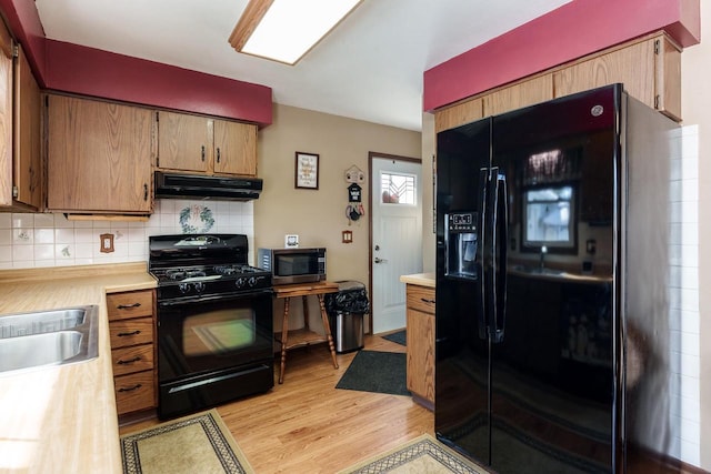 kitchen featuring brown cabinets, under cabinet range hood, light countertops, black appliances, and backsplash