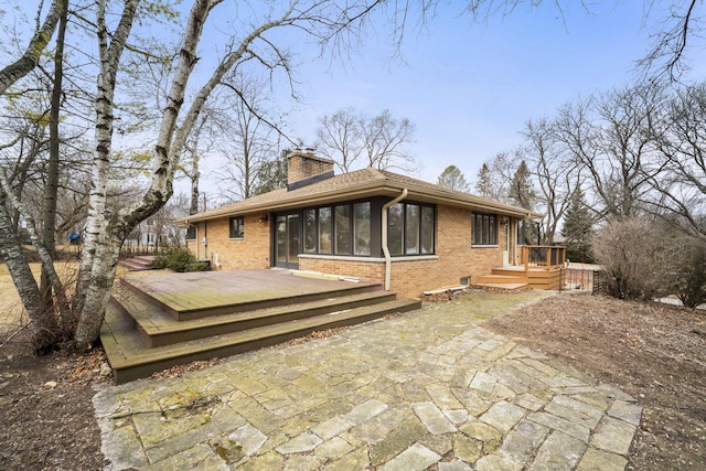 back of property featuring brick siding, a wooden deck, a chimney, a sunroom, and a patio area