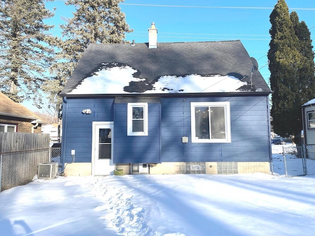 snow covered property featuring cooling unit, roof with shingles, fence, and a chimney