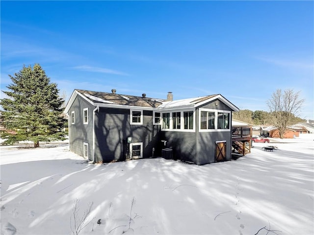 snow covered back of property with central AC, a chimney, and a sunroom