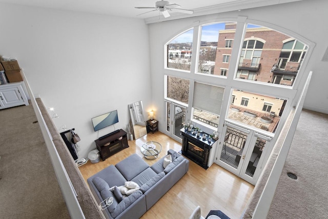 living room with a towering ceiling, a glass covered fireplace, wood finished floors, and a ceiling fan