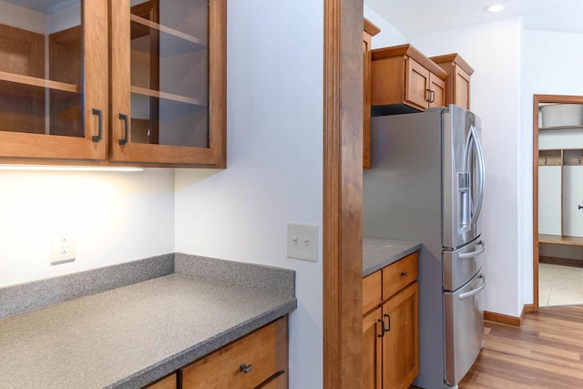 kitchen featuring stainless steel refrigerator with ice dispenser, light wood finished floors, dark countertops, brown cabinetry, and glass insert cabinets