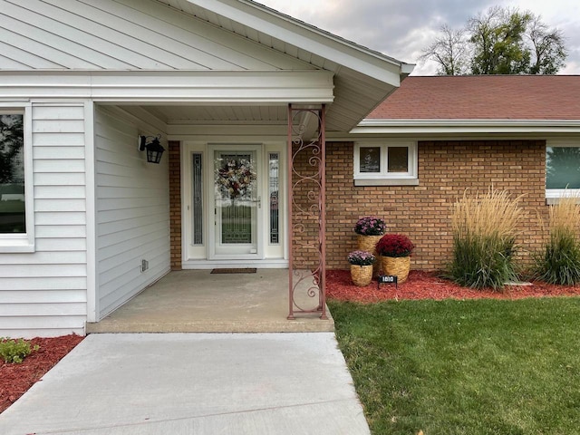 doorway to property featuring brick siding, a lawn, and a shingled roof