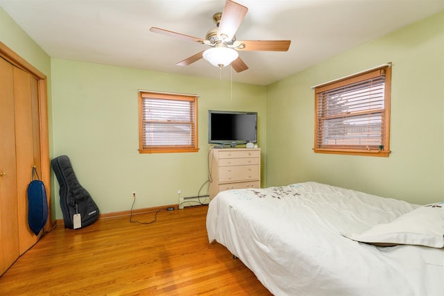 bedroom with light wood finished floors, a closet, a baseboard heating unit, a ceiling fan, and baseboards