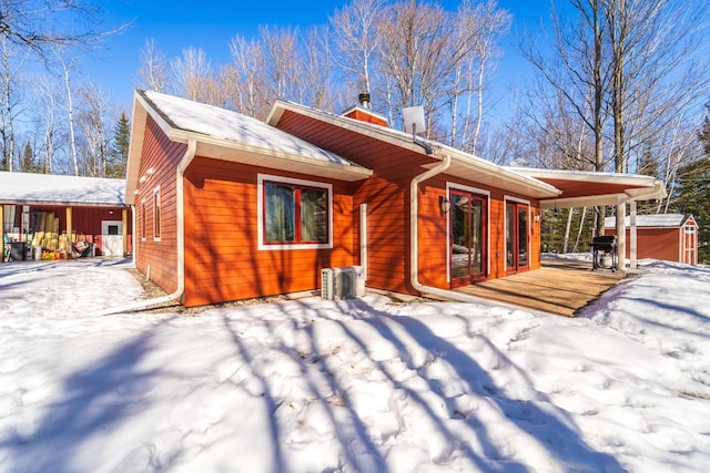 snow covered house featuring ac unit, a chimney, a storage unit, and an outbuilding