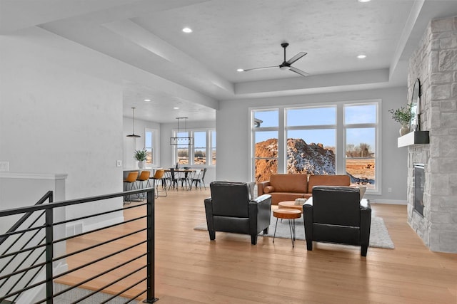 sitting room featuring a stone fireplace, a ceiling fan, baseboards, light wood-type flooring, and a raised ceiling