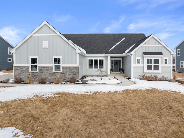 snow covered house featuring board and batten siding, a shingled roof, and stone siding