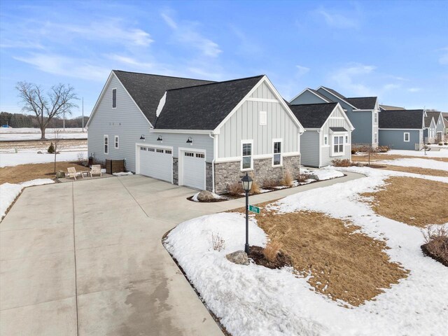 snow covered property with board and batten siding, a garage, concrete driveway, stone siding, and roof with shingles